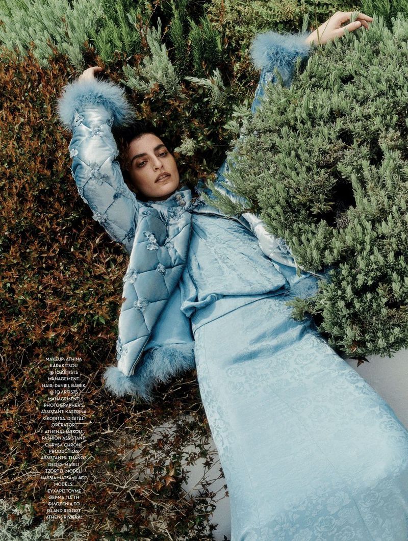 Woman in a blue jacket and dress poses lying in outdoor greenery. The portrait-orientation fashion photograph has bright, soft, diffused light.