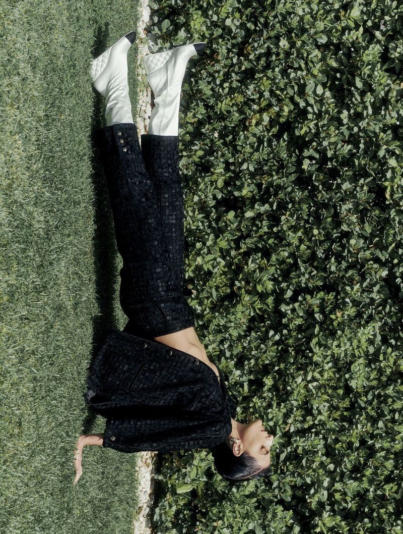 Fashion shot of a woman lying in a grassy field wearing a black jacket, black pants, and white boots, with a hedge background.