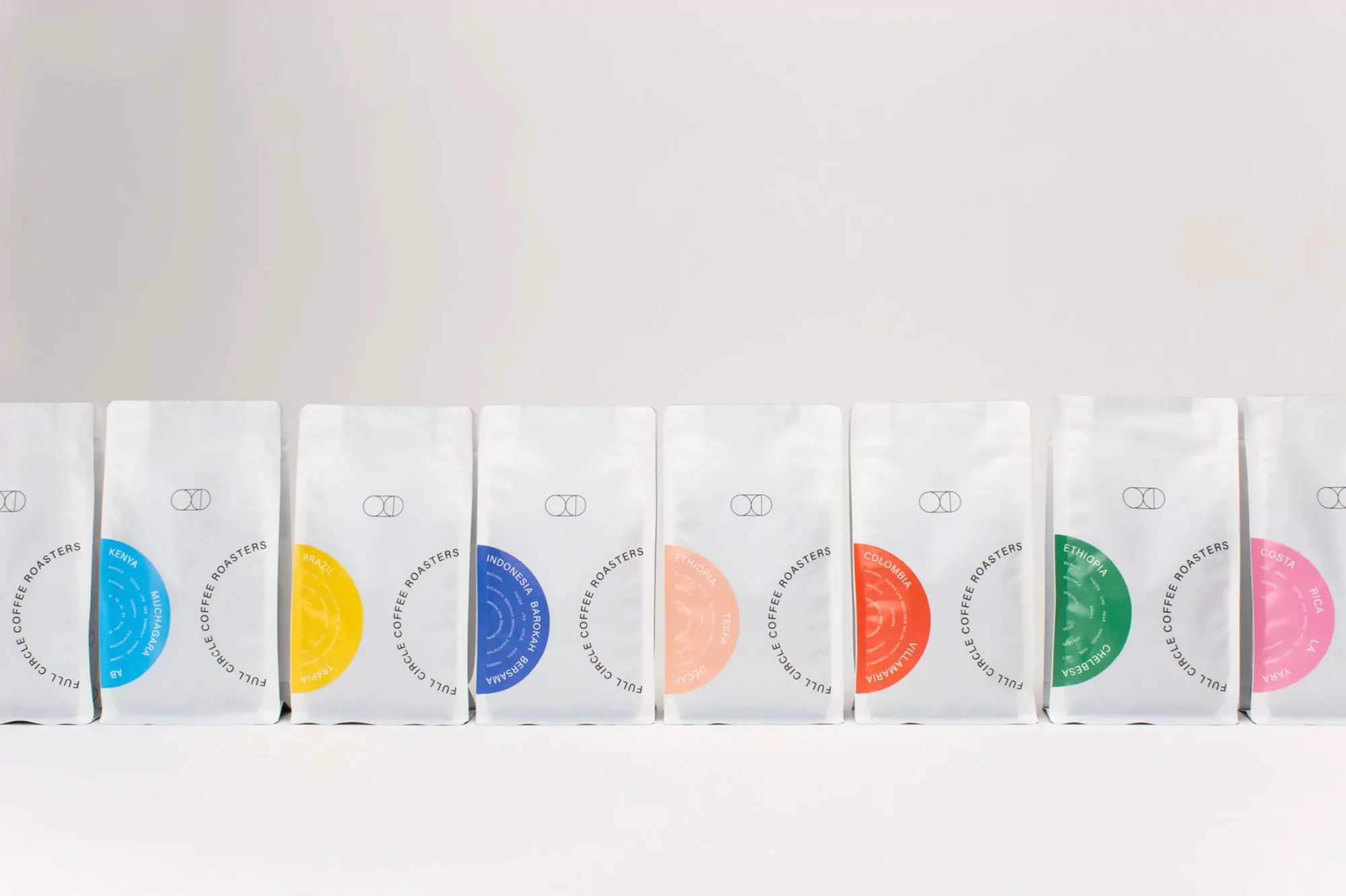 Seven white coffee bags from Full Circle Coffee Roasters lined up on a white surface, each labeled with a different origin and color.