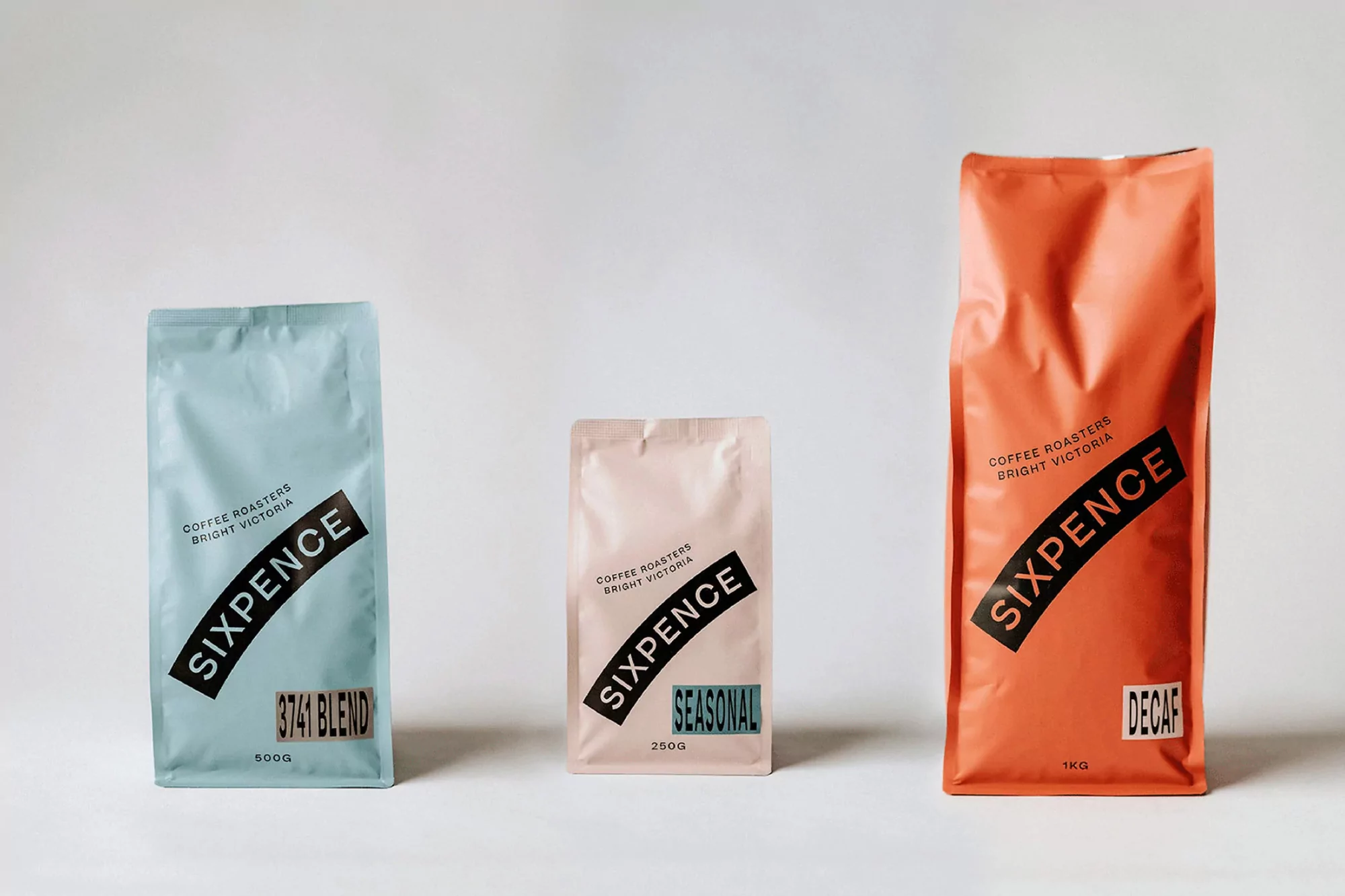 Three Sixpence coffee bags in blue, pink, and orange colors stand against a white background, displaying a clean and modern design.