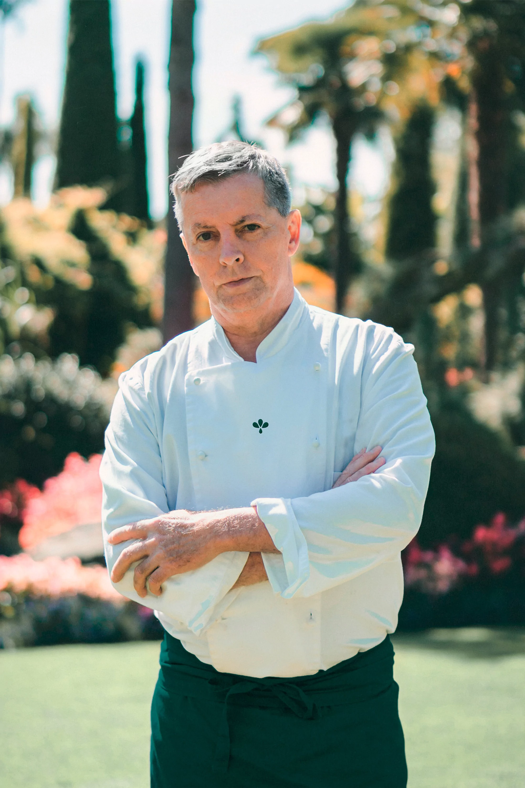 Portrait of a confident male chef in a white uniform with arms crossed, standing outside in a green garden setting on a sunny day.