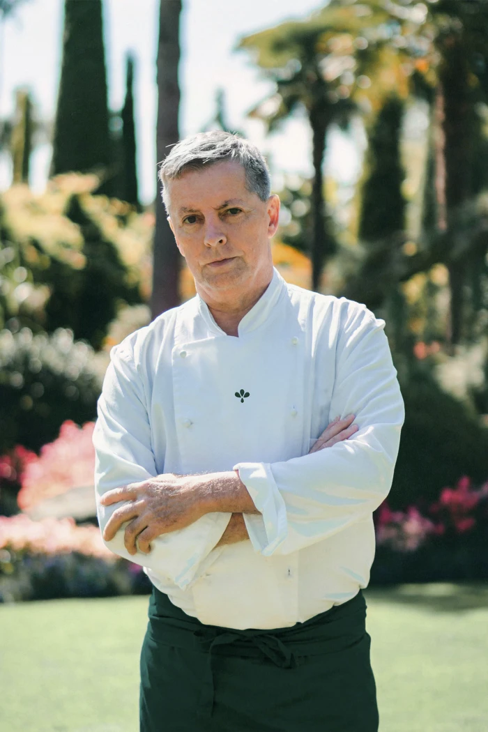 Middle-aged man in a white chef coat with arms crossed standing outdoors in a garden setting for Flor & Fjære brand identity.