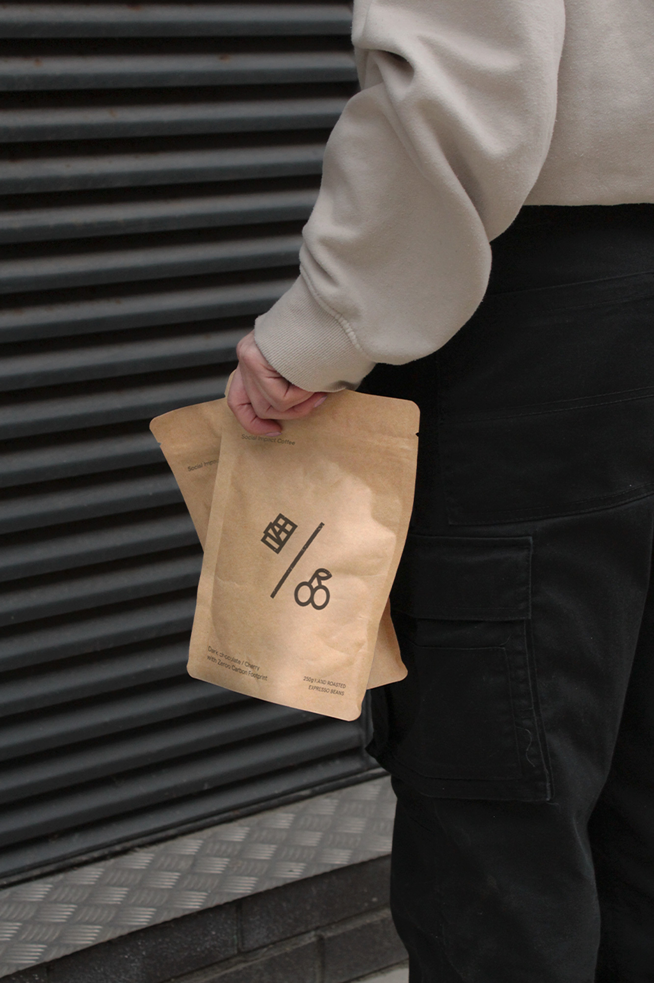 Unidentifiable person holding two brown paper bags with printed logos against a dark grey ridged wall in an outdoor setting.