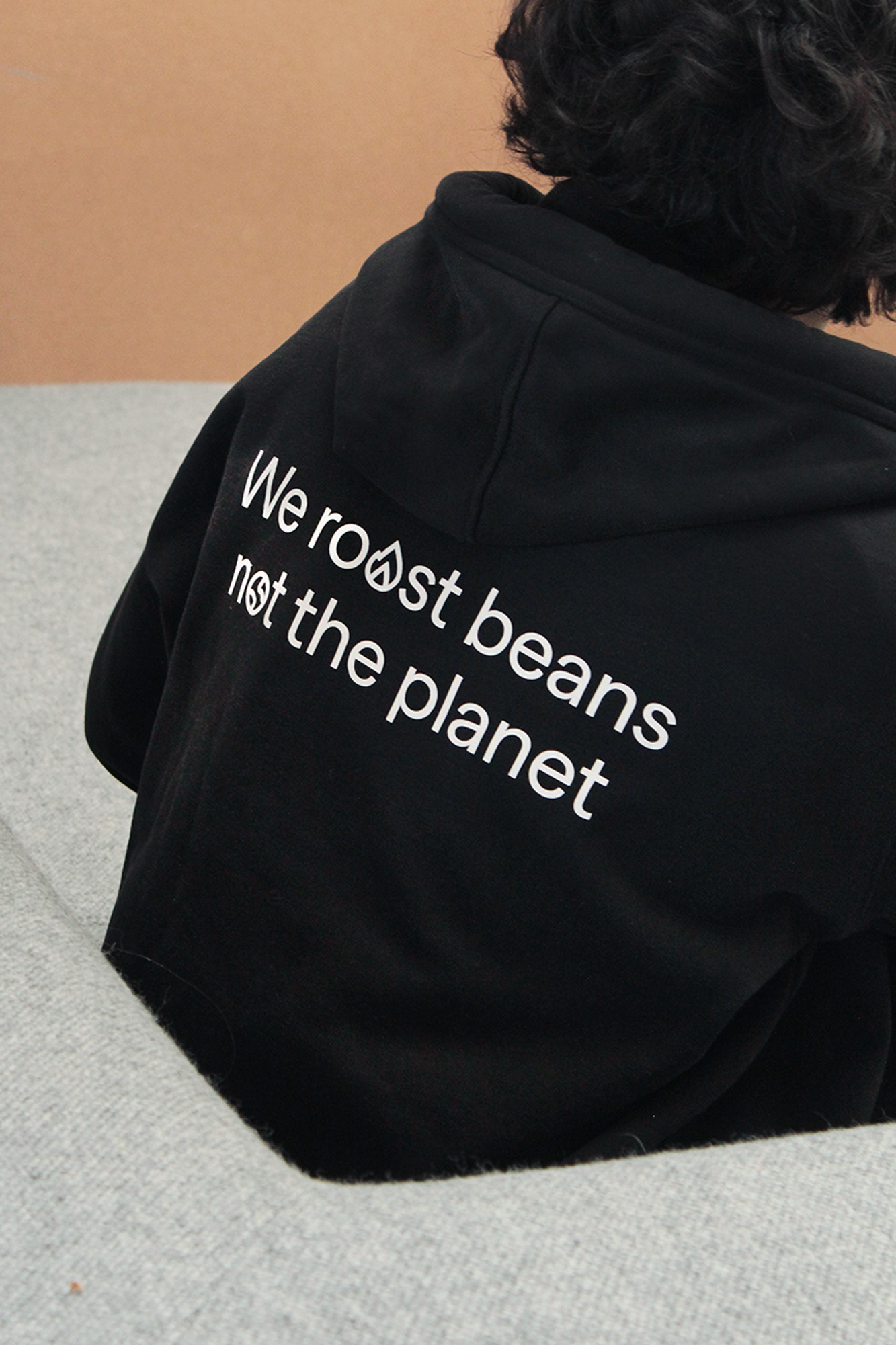 Person wearing a black hooded sweatshirt with the text "We roast beans not the planet" in white on the back, sitting on a grey couch.