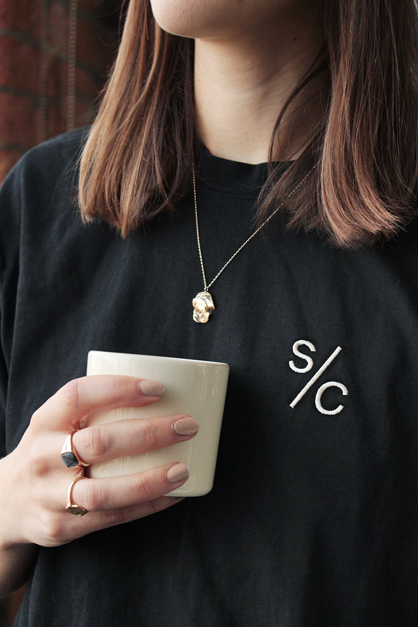 Brunette woman wearing black t-shirt and holding white mug, gold necklace, and rings with a brick wall in the background.