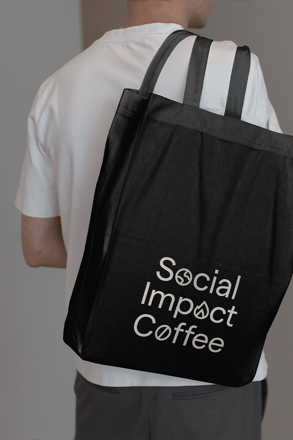 Person wearing a white t-shirt carries a black tote bag with white Social Impact Coffee branding against a neutral background.