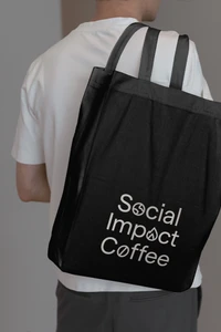 Person wearing a white t-shirt carries a black tote bag with white Social Impact Coffee branding against a neutral background.