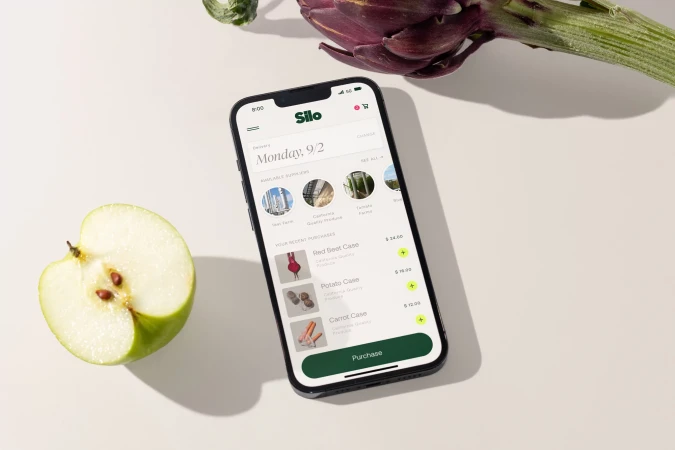 A smartphone displaying a grocery shopping app, with an apple half and artichoke, showcasing food and technology integration on a table.