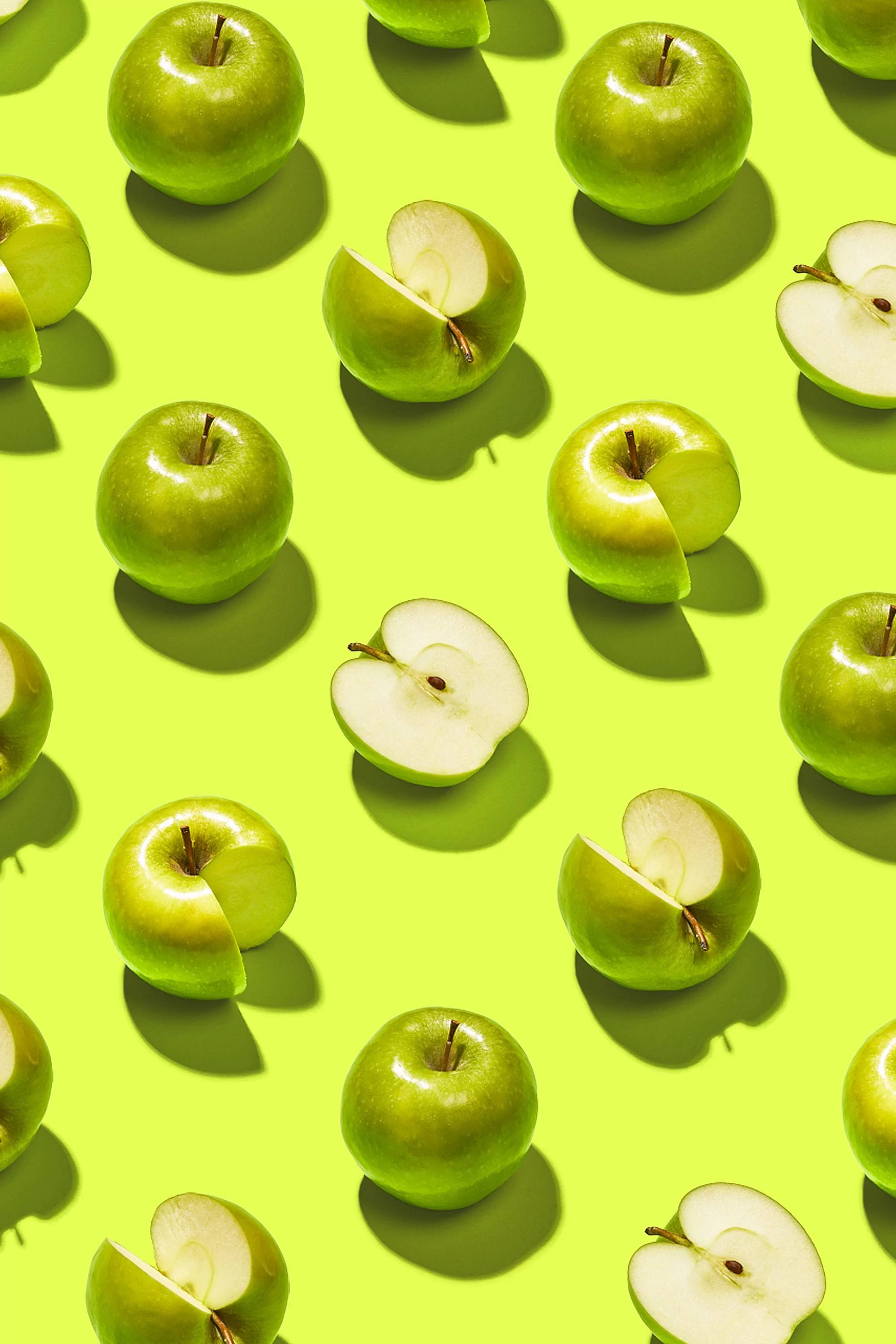Overhead shot of a repeating pattern of whole and sliced green apples on a bright chartreuse background with contrasting hard shadows.
