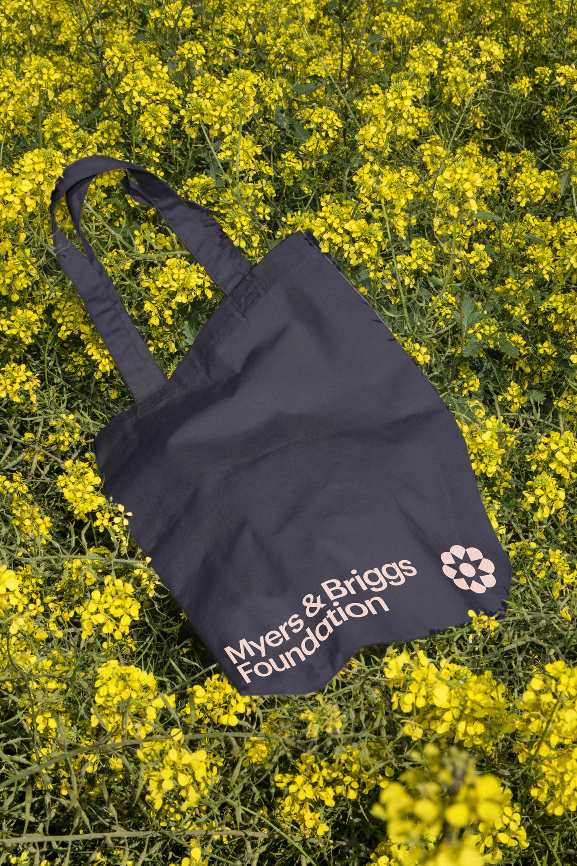 Black tote bag with Myers & Briggs Foundation logo sits in a field of yellow flowers, creating a striking contrast and eco-friendly vibe.