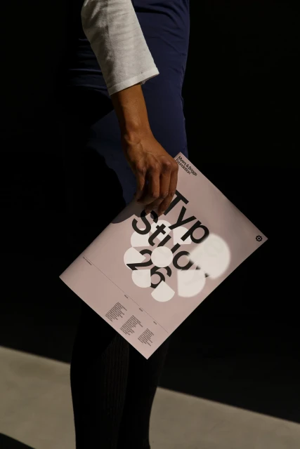 Adult holding a light pink magazine with white and black typography. Dark background, light and shadow.