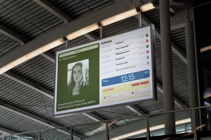 Digital signage displays Dr. Marisa Koonz and a schedule with times, live feed, and green and white design in an indoor setting.