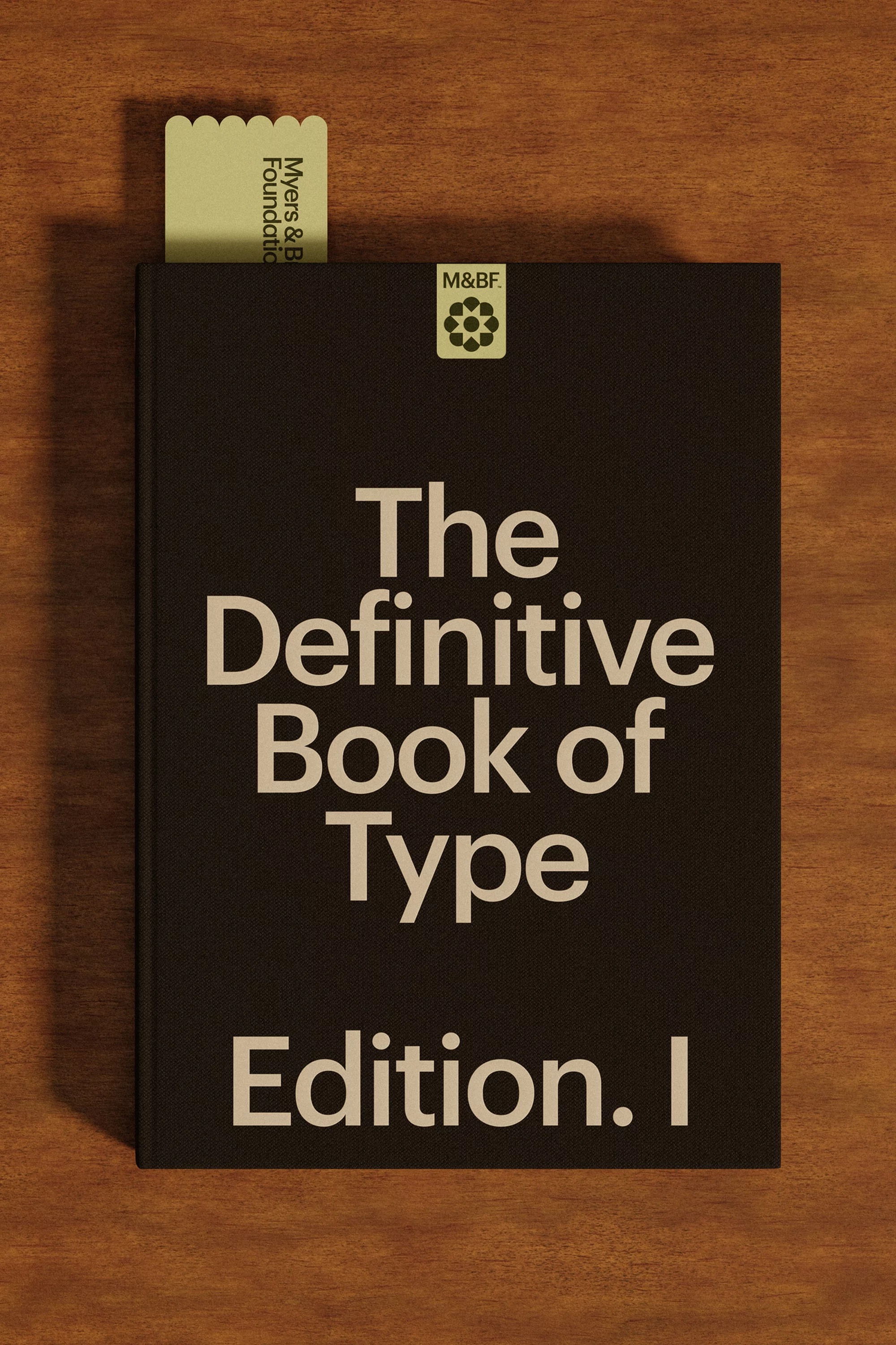 A black hardcover book titled 'The Definitive Book of Type, Edition I' sits on a wood surface, accompanied by a light green bookmark.
