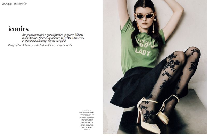 A young woman wearing a green T-shirt, black skirt, patterned tights, and high heels sits on a gray surface in a fashion shot.