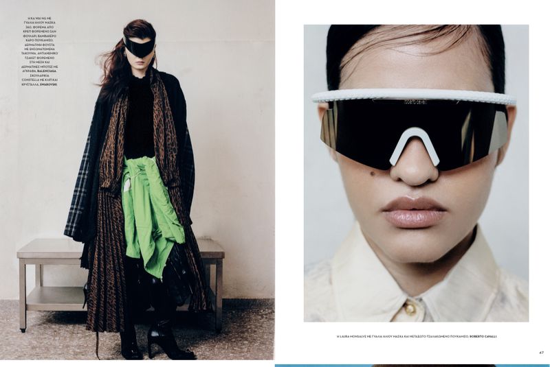 Editorial fashion shot with a woman wearing an avant-garde mask, layered coats, and boots, posing against a muted wall and metal bench.