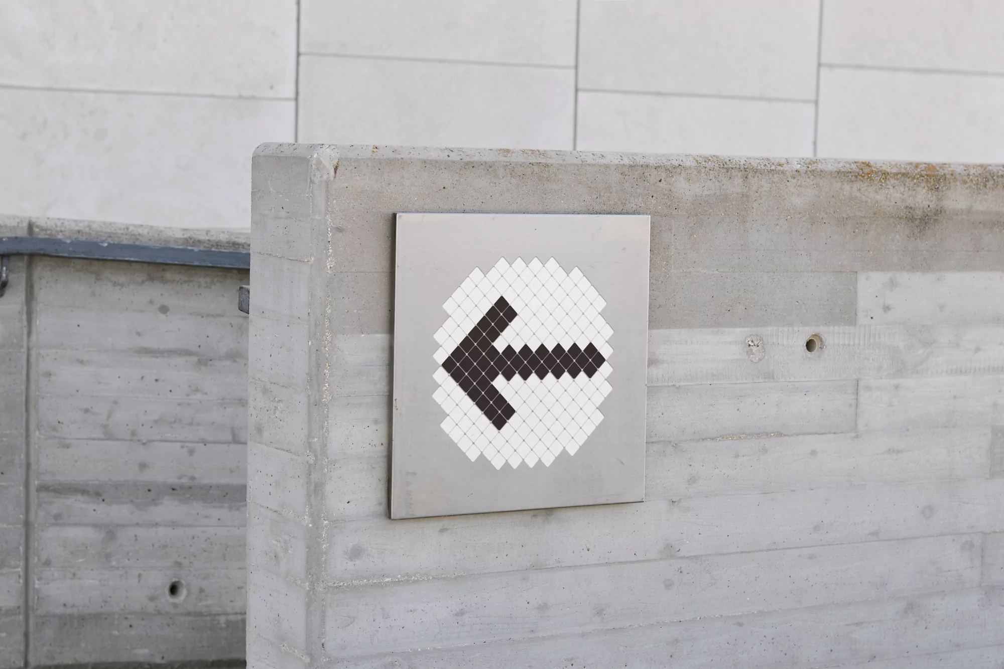 Directional sign with a black arrow pointing left on a silver square, mounted on a concrete wall under natural daylight.