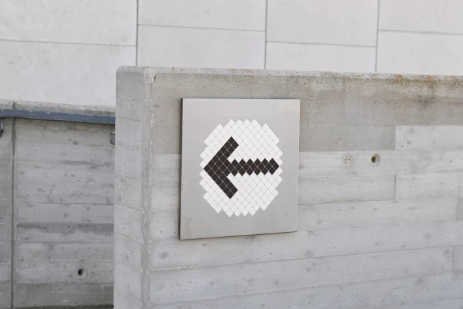 Directional sign with a black arrow pointing left on a silver square, mounted on a concrete wall under natural daylight.