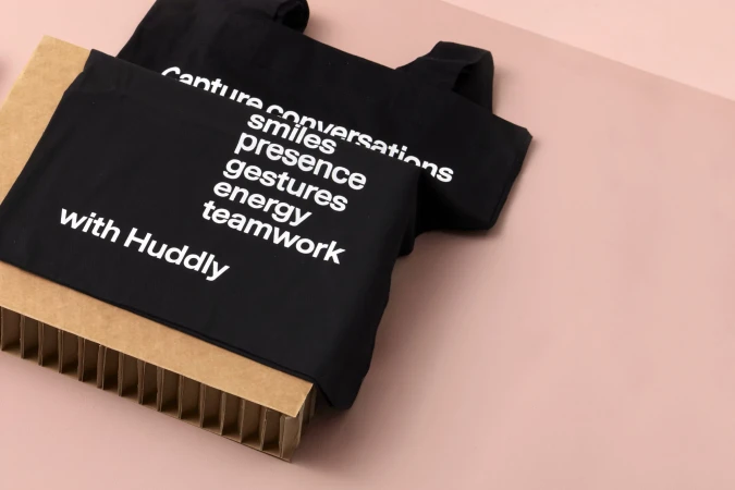 Two black tote bags featuring white text, 'Capture conversations, smiles, presence, gestures, energy, teamwork with Huddly,' on a pink background.