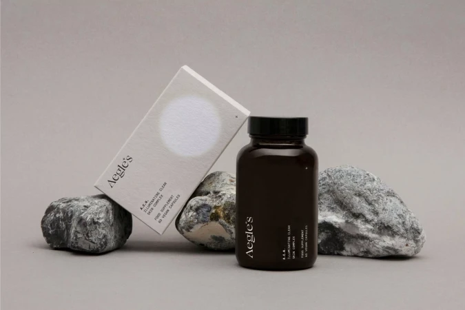 Brown supplement bottle and product box with Aegle's logo displayed on gray rocks against a grey background in a studio shot.