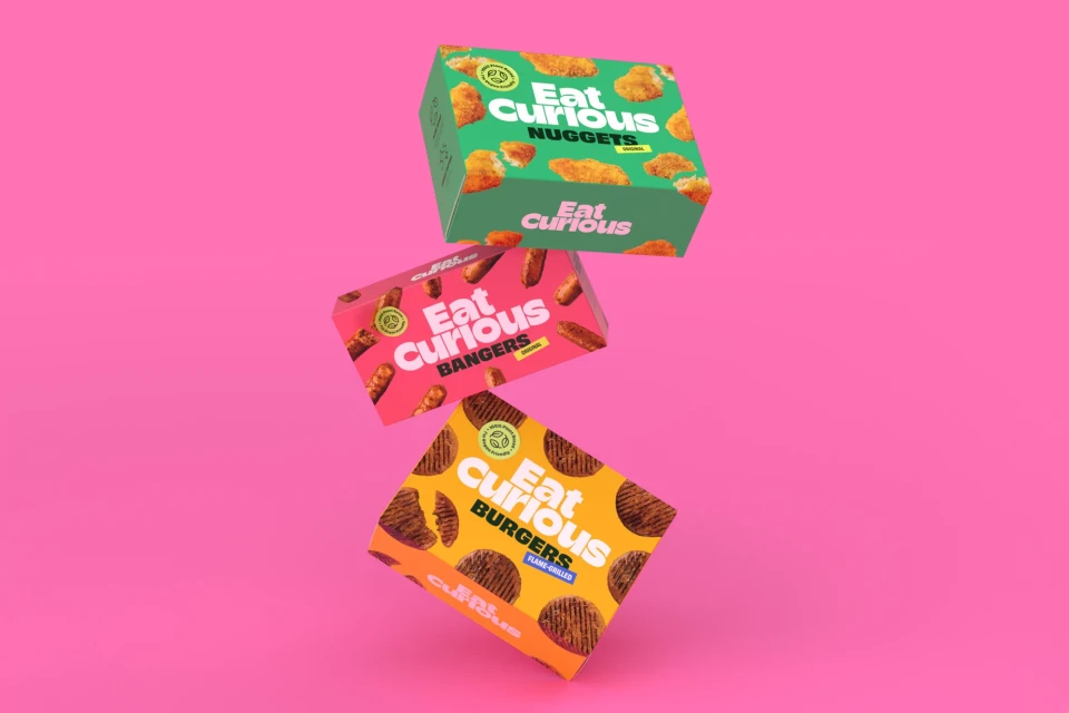 Three colorful product boxes from Eat Curious balanced on each other against a bright pink background in a studio product shot.
