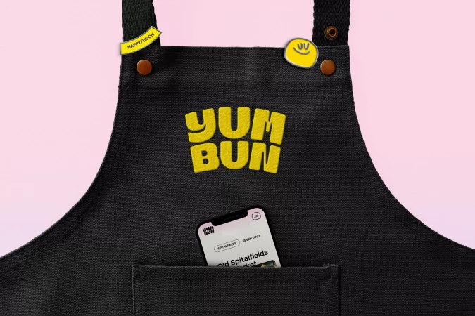 Black apron with the yellow Yum Bun logo and a cell phone in the pocket against a pink background, product promotion.