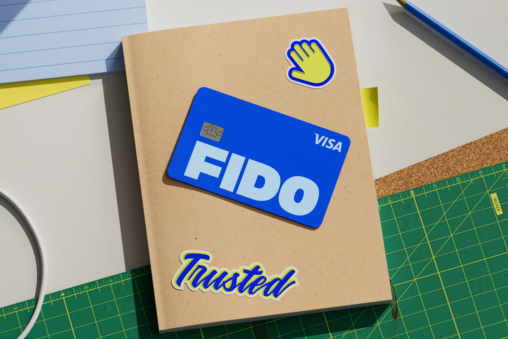 A blue Fido Visa credit card and a hand sticker on a beige notebook, set against a green cutting mat.