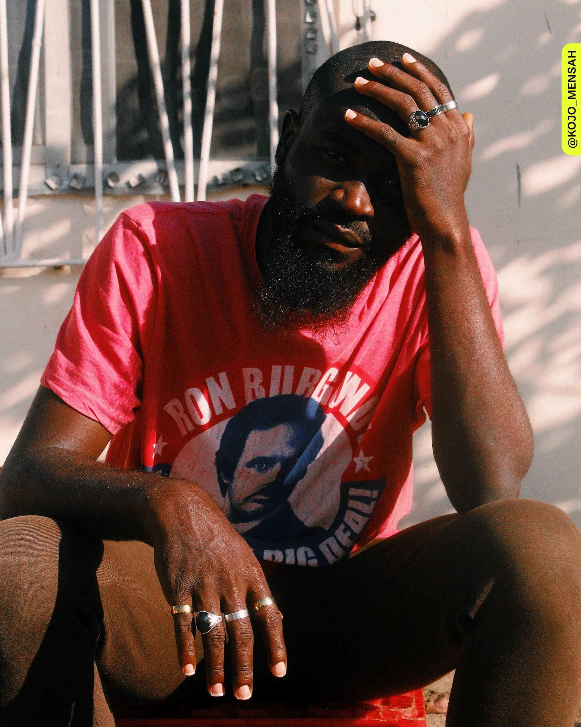 Bearded man in a 'Ron Burgandy' shirt sitting outdoors on a red stool with sunlight and shadows on his face, body, and background.