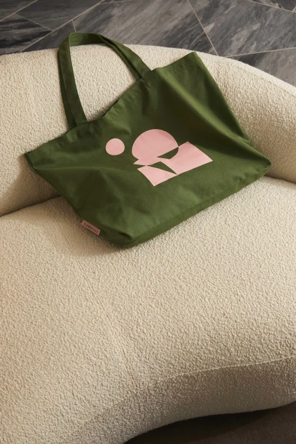 Olive green tote bag with pink graphic logo on a textured beige couch. Fashionable and sustainable accessory for everyday use.
