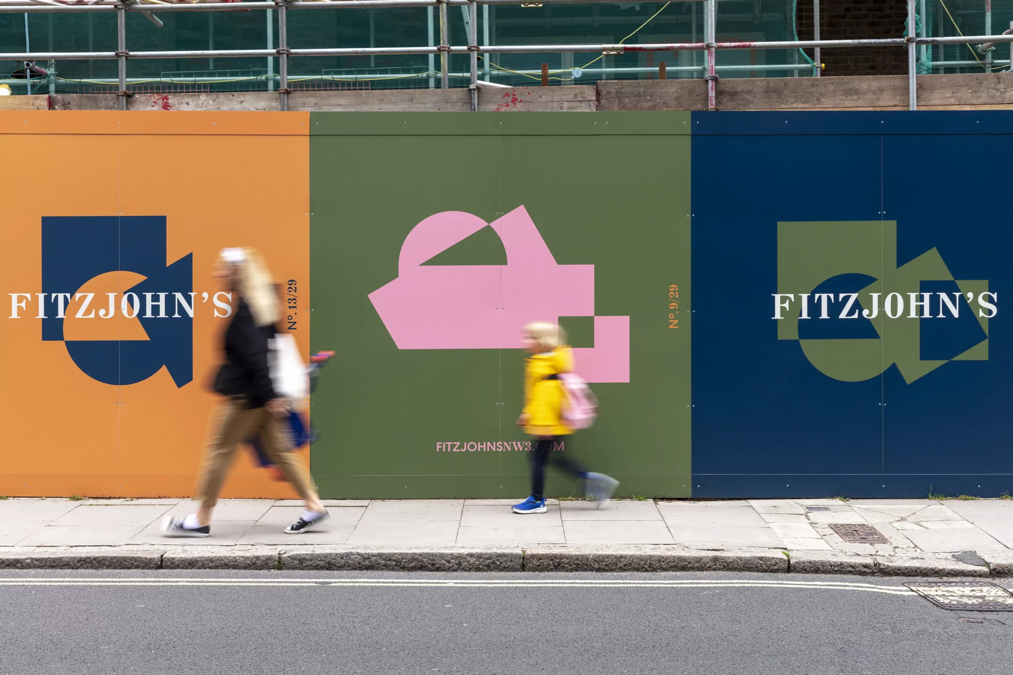 Two blurred figures walk along a sidewalk in front of an abstract graphic mural in shades of orange, green, and blue.