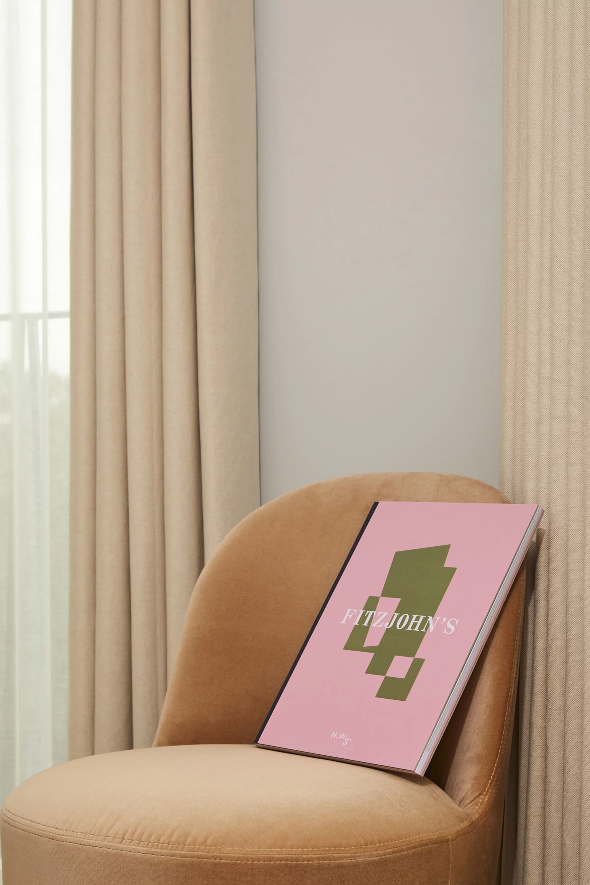 Pink book with green geometric shapes on a tan velvet chair near beige drapes and a light gray wall in an indoor setting.