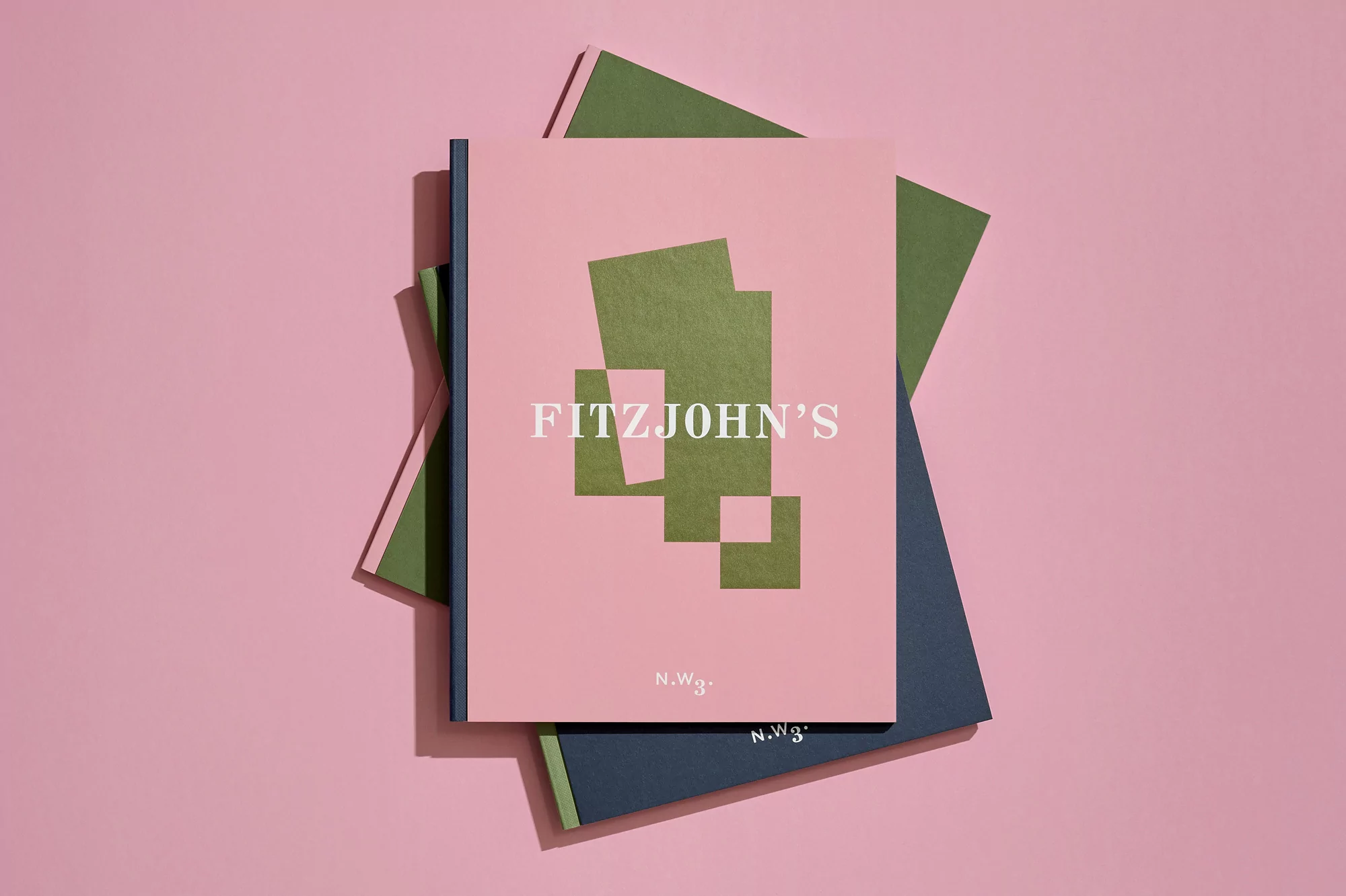 Flat lay of a pink book with an abstract geometric design and text, stacked on top of two other books against a pink backdrop.
