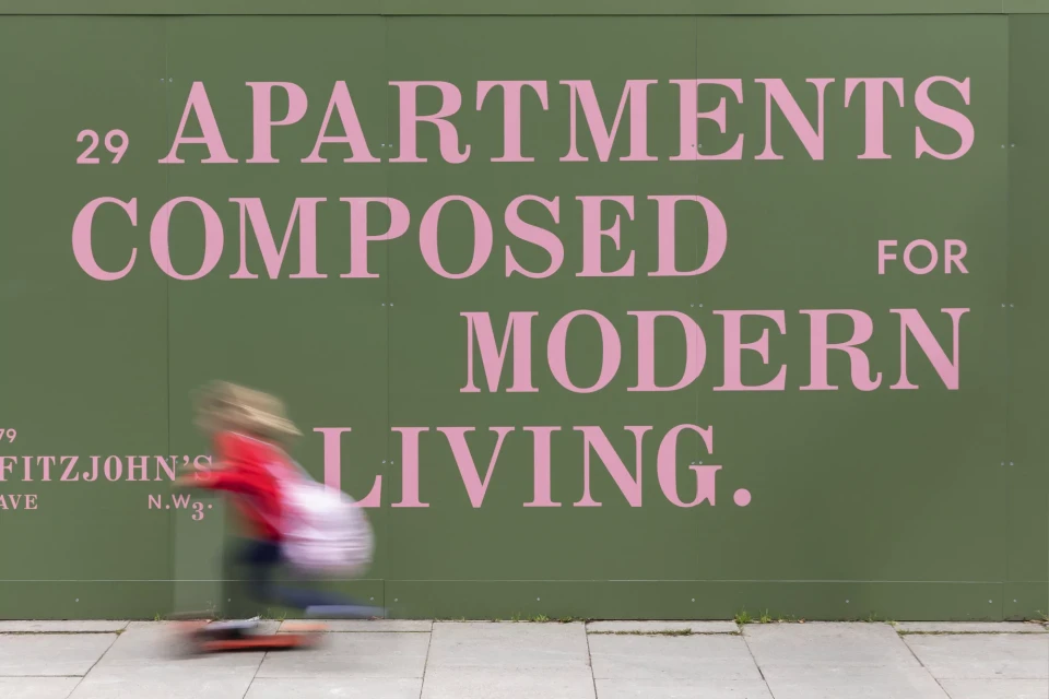 Blurred motion of a child skateboarding on sidewalk in front of green wall with pink apartment advertisement in daylight.