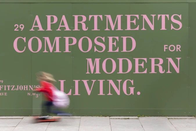 Blurred motion of a child skateboarding on sidewalk in front of green wall with pink apartment advertisement in daylight.
