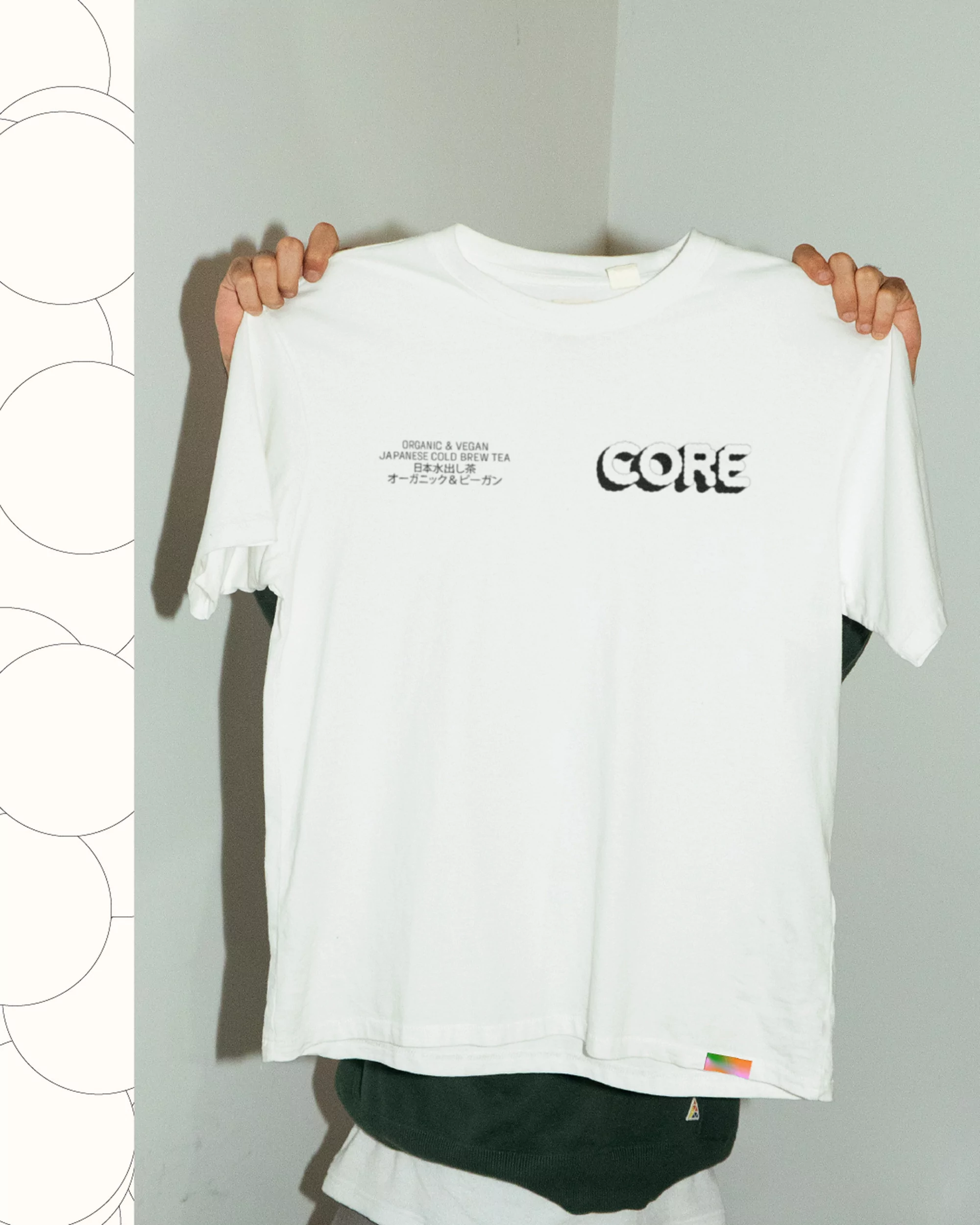 A white t-shirt with 'CORE' text held up by two hands against a grey and white wall, capturing a minimalist, casual fashion aesthetic.