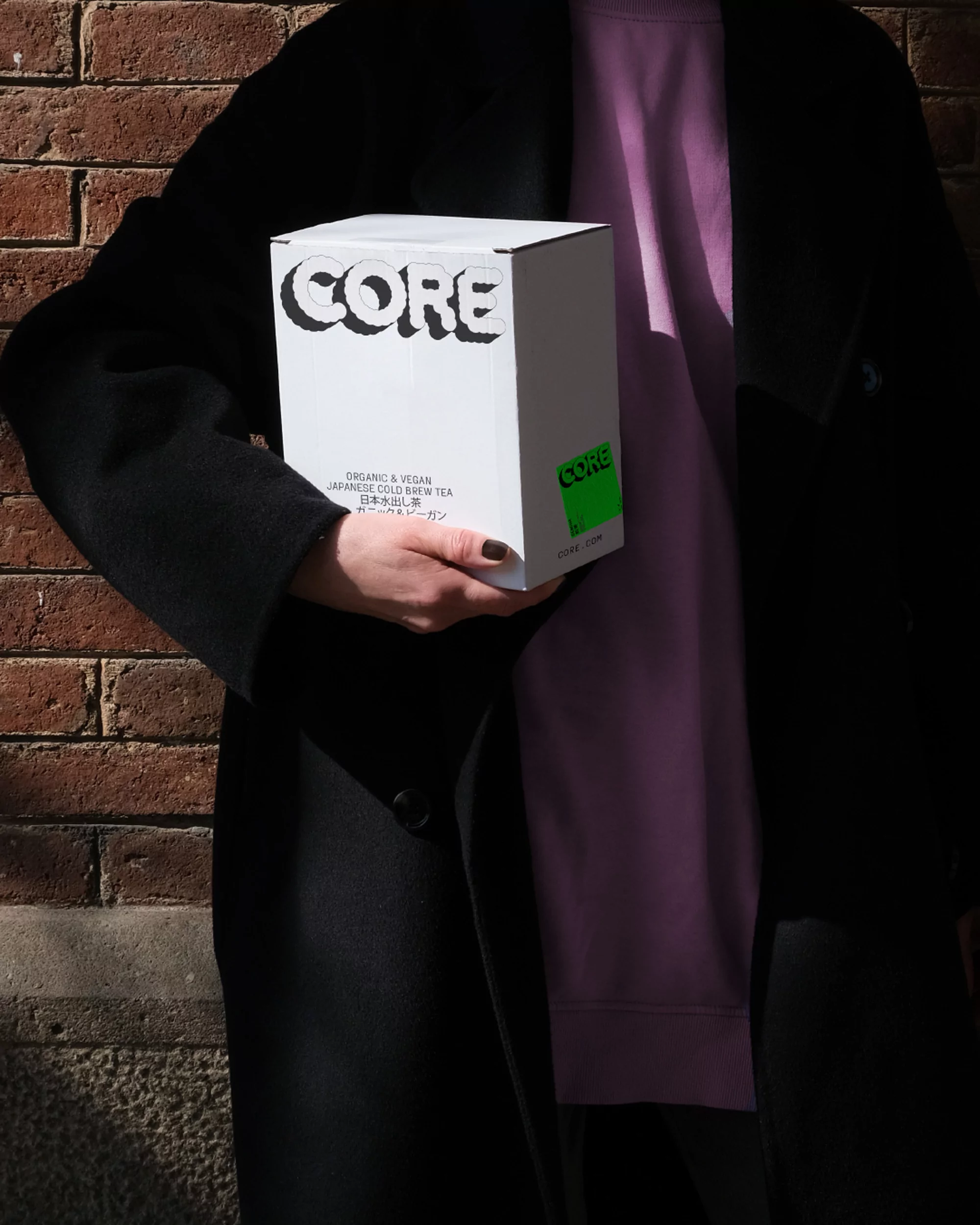 Person holding a white Core brand beverage box in front of a brick wall, wearing a black coat and lilac sweatshirt. Product shot.