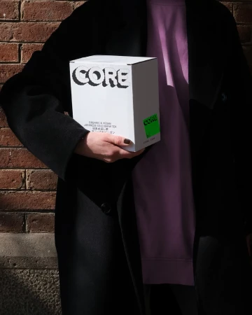 Person holding a white Core brand beverage box in front of a brick wall, wearing a black coat and lilac sweatshirt. Product shot.