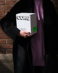 Person in a black coat holding a white CORE branded box against a brick wall