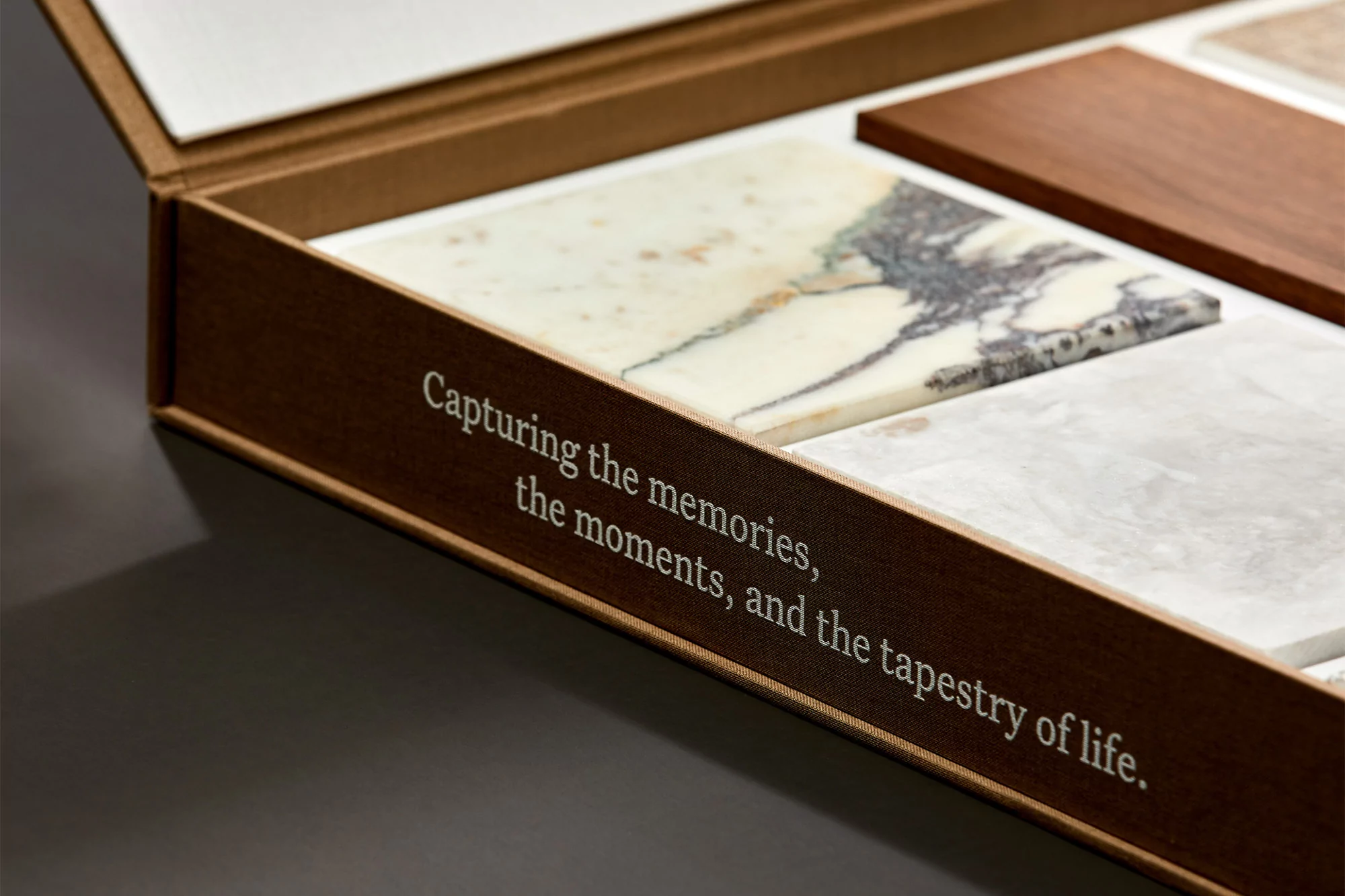 Close-up of an open, brown box containing marble and wood sample tiles. The box reads, 'Capturing the memories, the moments, and the tapestry of life.'