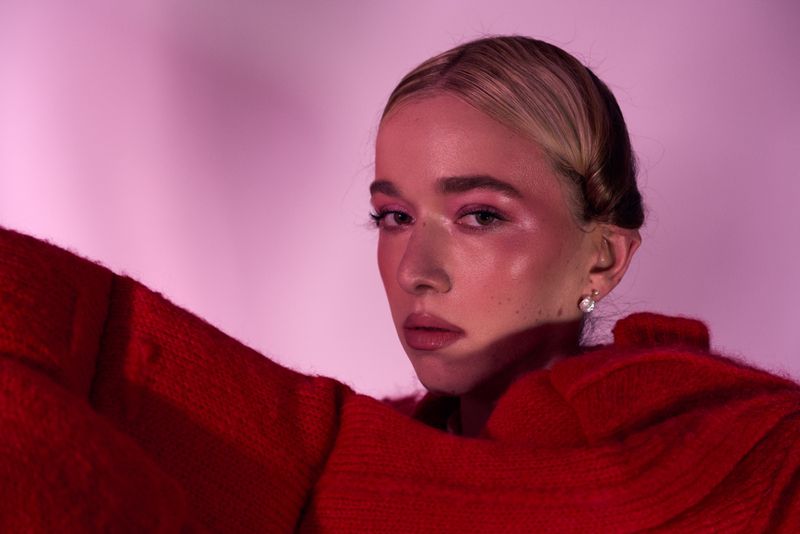 Close-up portrait of a young woman with fair skin and blonde hair wearing a red sweater against a pink background in a studio setting.