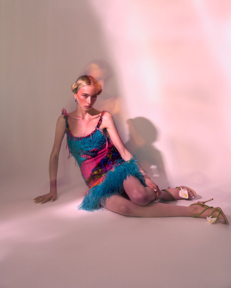 Fashion portrait of a woman with an elegant updo, wearing a colorful mini dress with feather trim, sitting against a neutral backdrop.