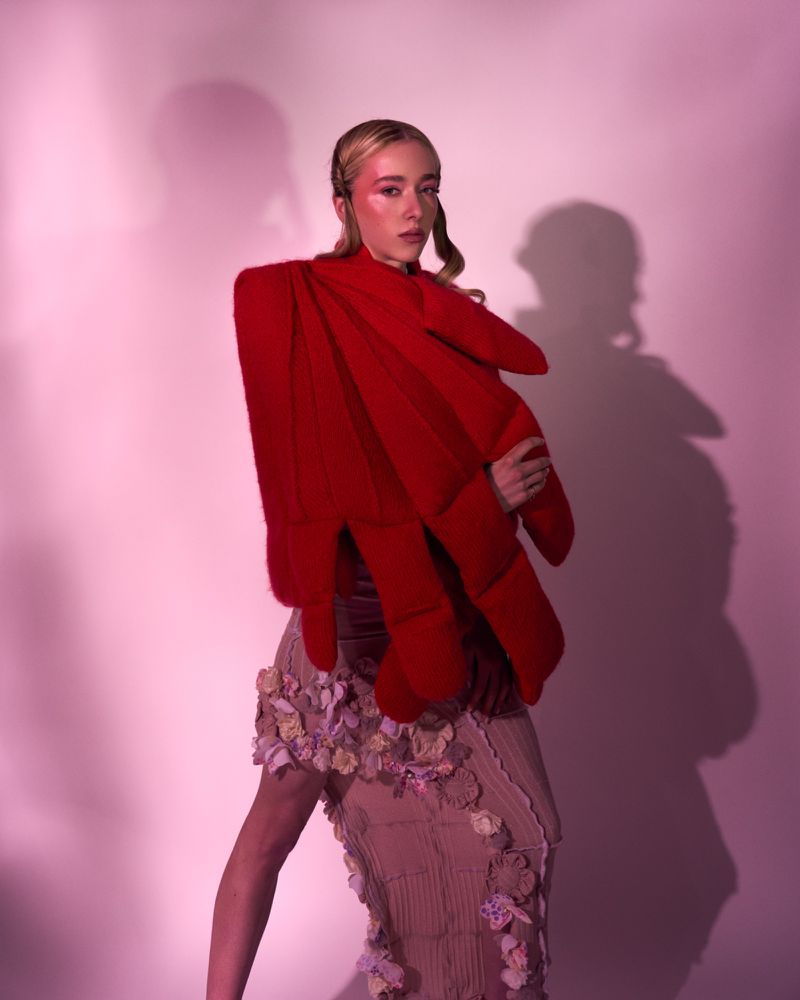 Woman poses in a red sculptural jacket and beige floral skirt in a studio with pink lighting, conveying avant-garde fashion.