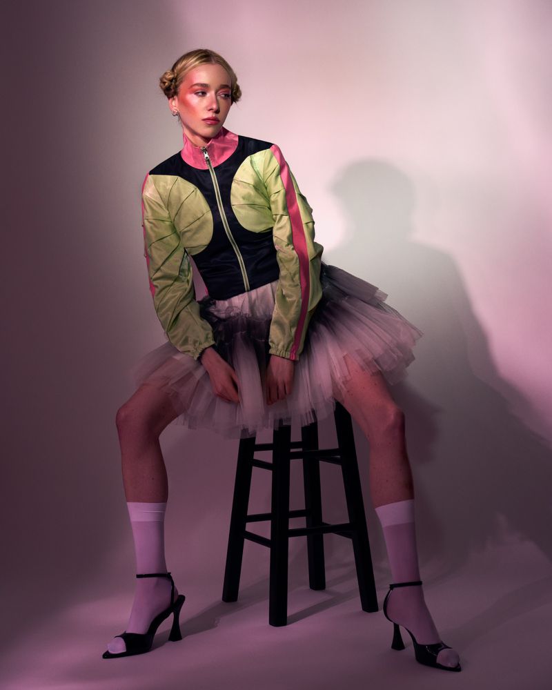 A woman with blonde hair in a jacket and tutu poses on a stool in a photo studio with pink and purple lighting, showcasing a creative portrait.