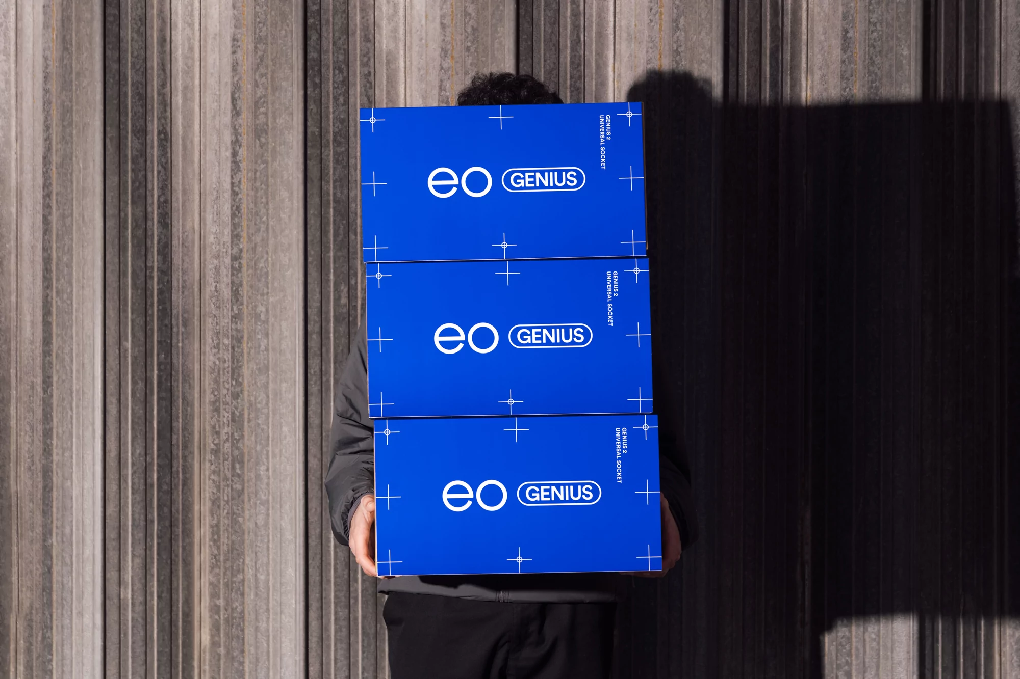 Person carrying a stack of three blue boxes with the words 'eo GENIUS' printed on them, against a textured gray background.