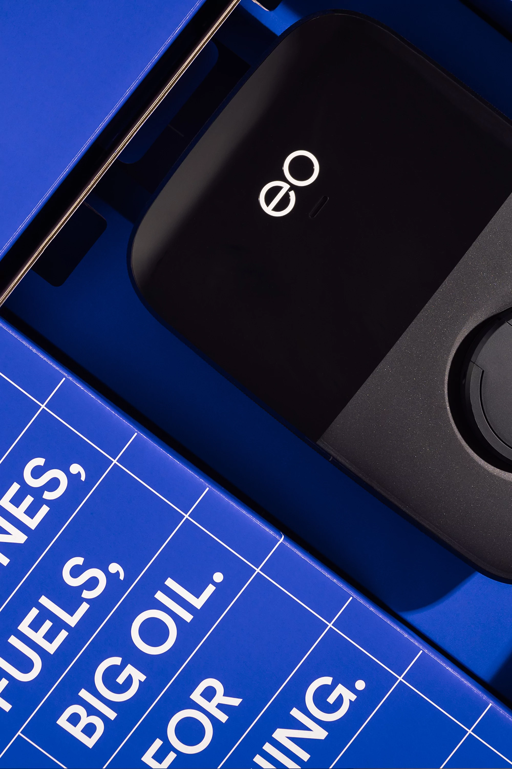 Close-up of a sleek black device with the 'eo' logo, nestled in a blue box with white grid lines and the text 'FUELS, BIG OIL'