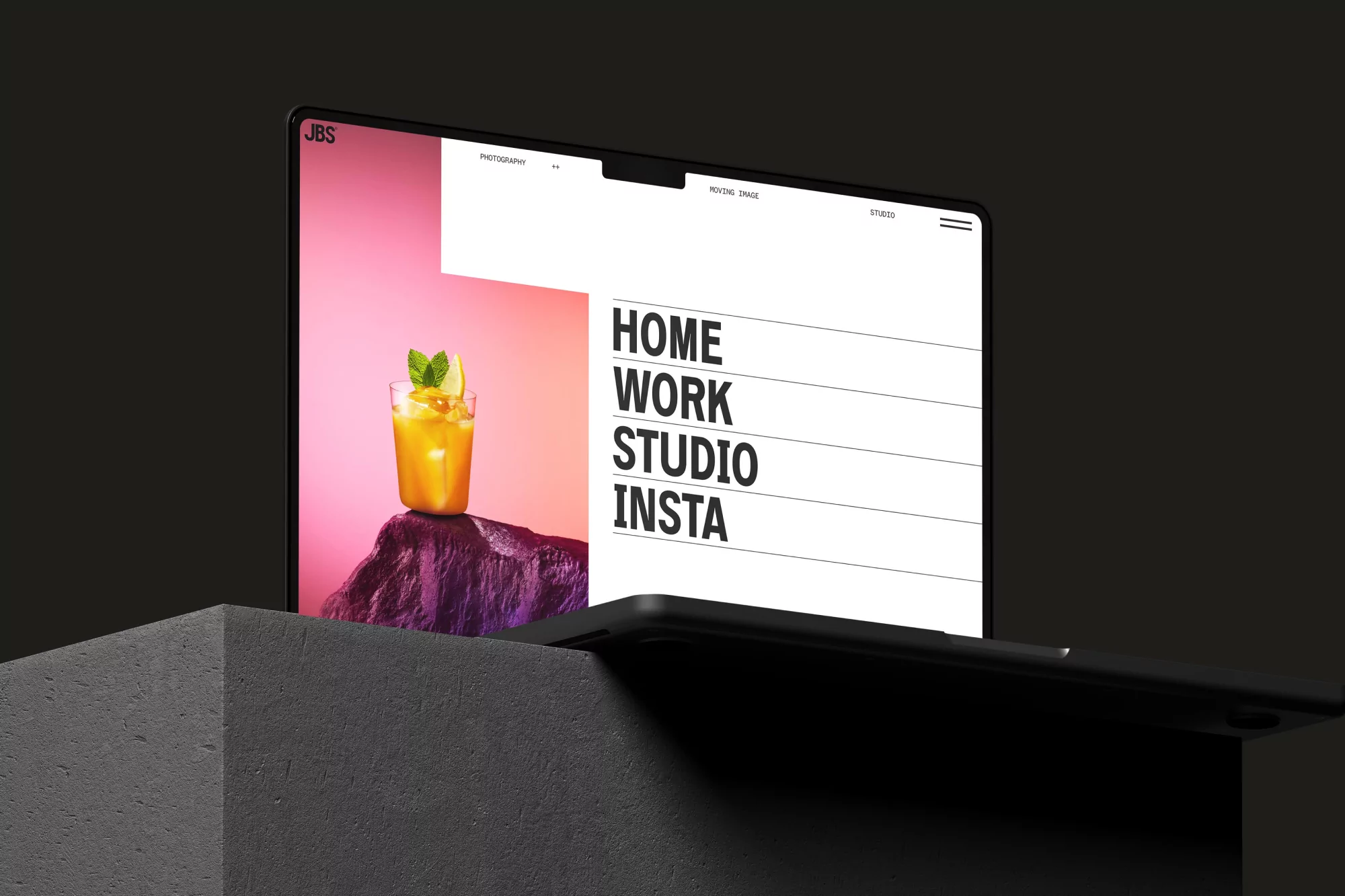 Laptop displays website featuring a glass of orange drink with lime and mint, set against pink backdrop, atop dark rock surface.
