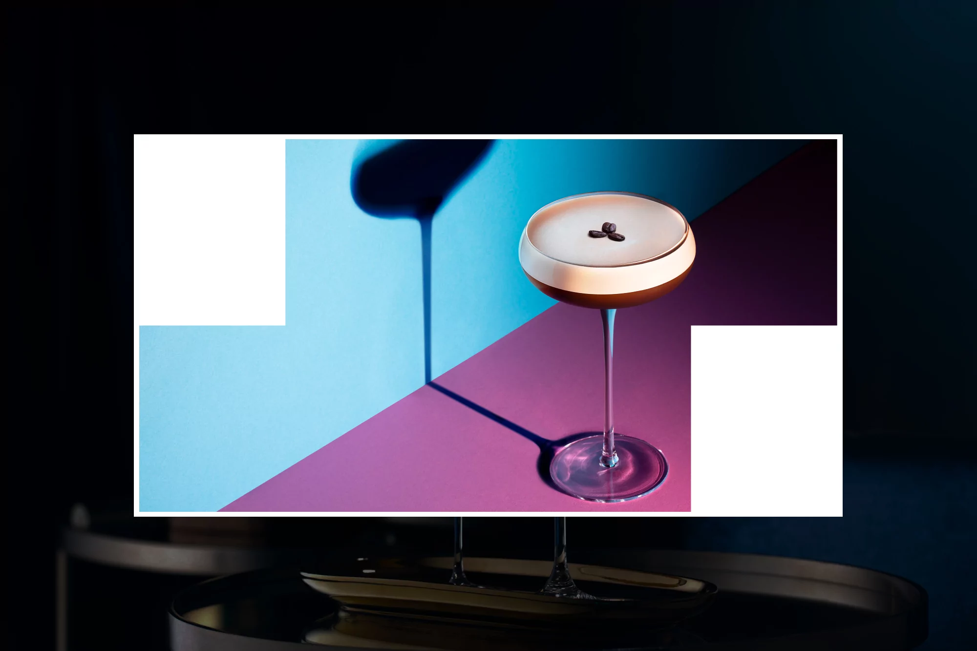 A dark cocktail in a clear stemmed glass sits on a table against a split light blue and purple background with dark shadows.
