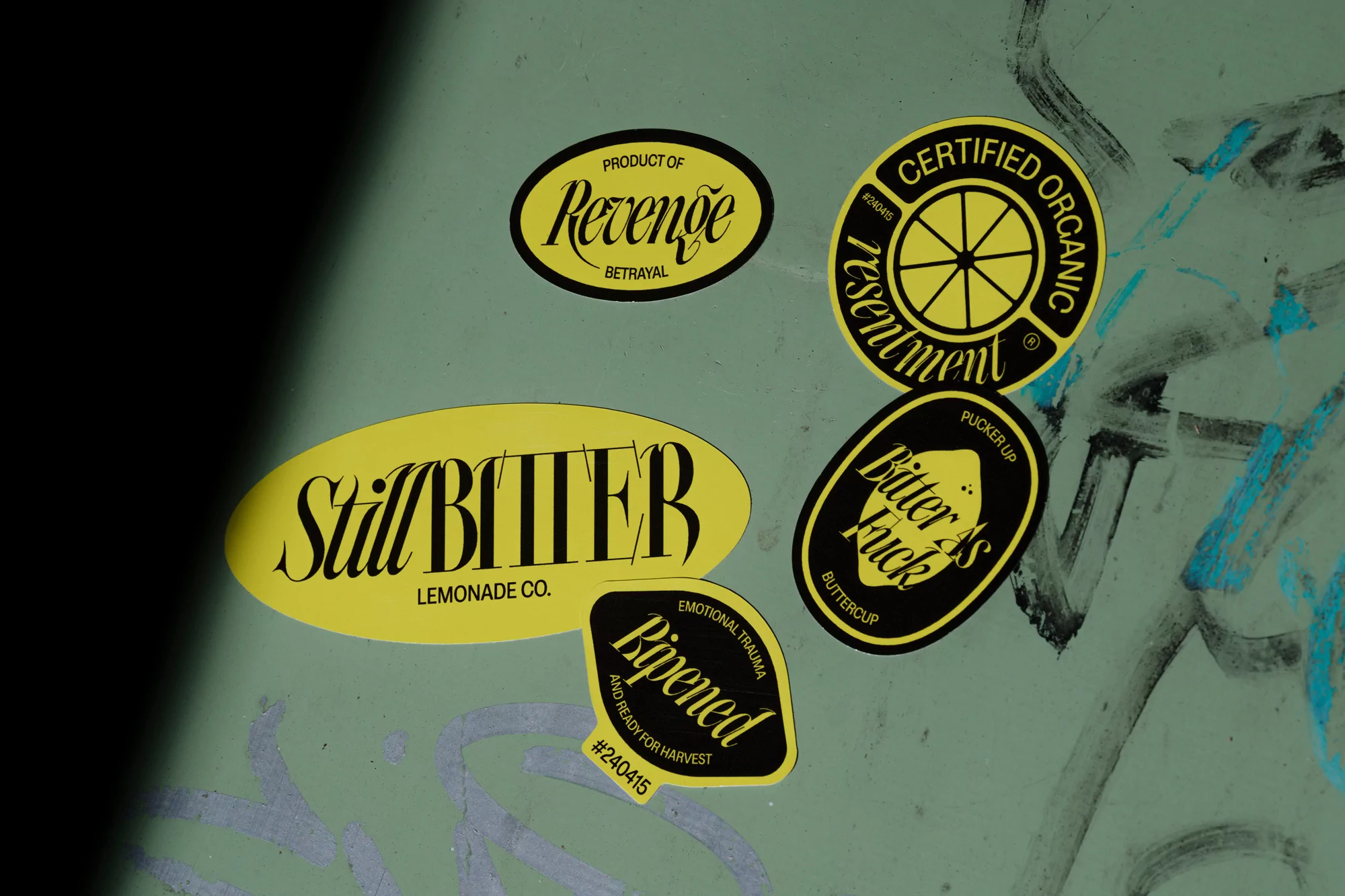 Close-up of colorful, stylized stickers with text such as 'Certified Organic,' 'Revenge,' and 'Still Bitter' on a green surface.