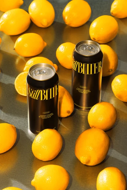 Two black and yellow cans of Still Bitter lager surrounded by bright yellow lemons on a reflective silver surface.