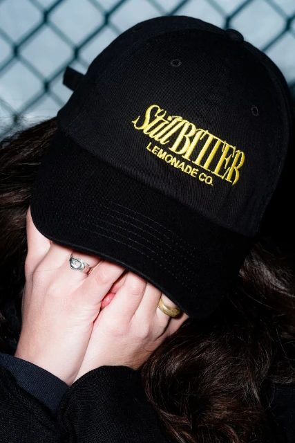 Young adult woman with dark hair wearing a black baseball cap with yellow embroidery covering her face with her hands.