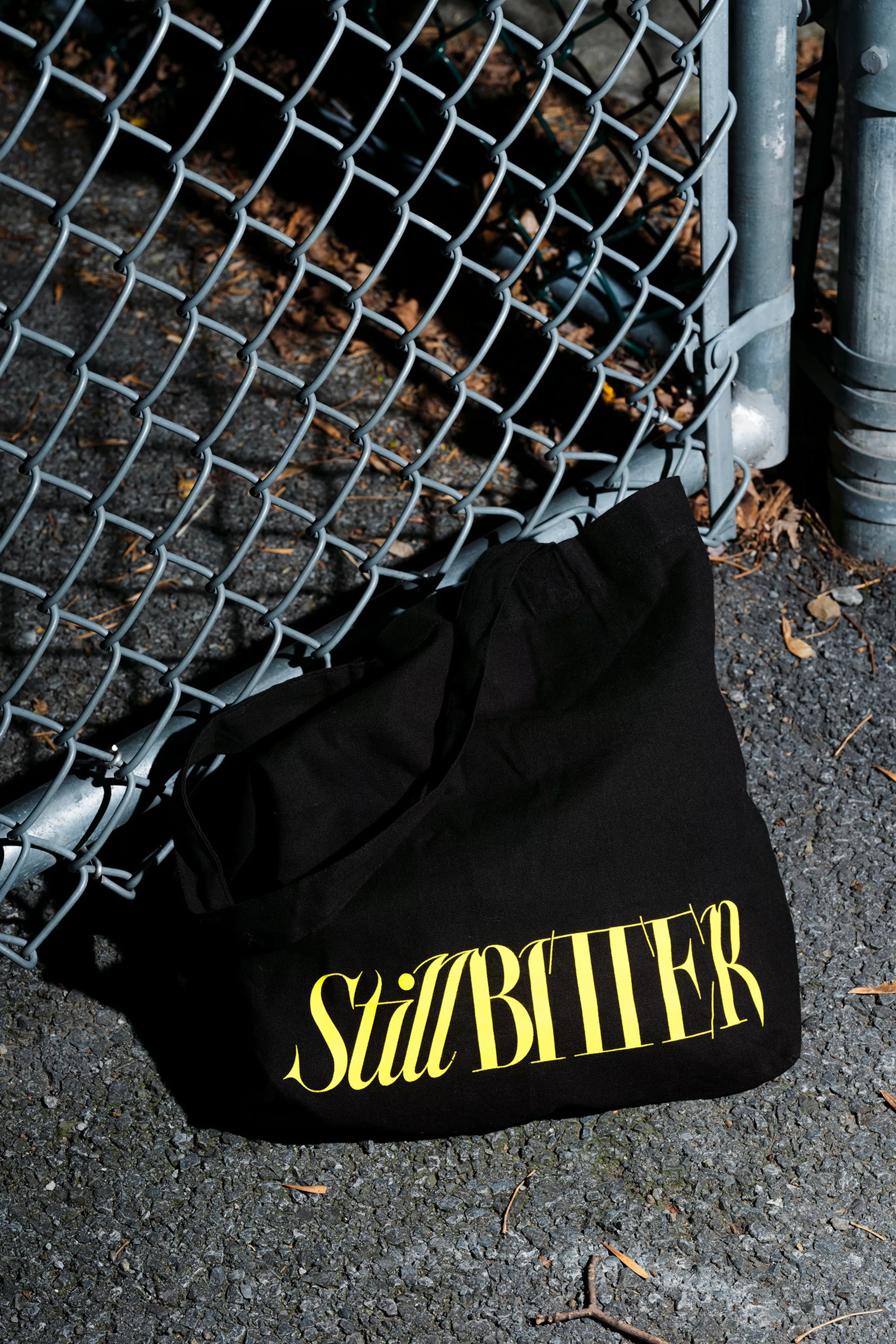 A black tote bag with yellow text rests against a chain-link fence on cracked asphalt outdoors, casting a shadow on a sunny day.